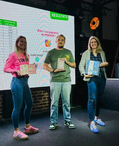 Trois personnes debout sur une scène, chacune tenant un coffret, devant un écran affichant « RÉSULTATS » et « Bravo aux gagnants du PODIUM ! ».
